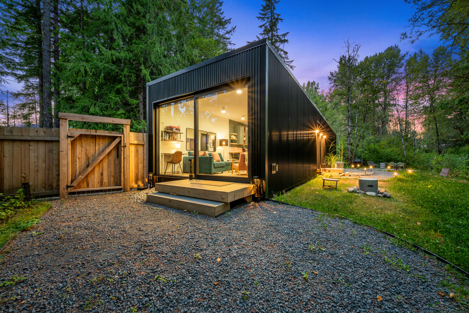 The guest house at twilight — glass wall glowing, fenced yard, fire pit, forest