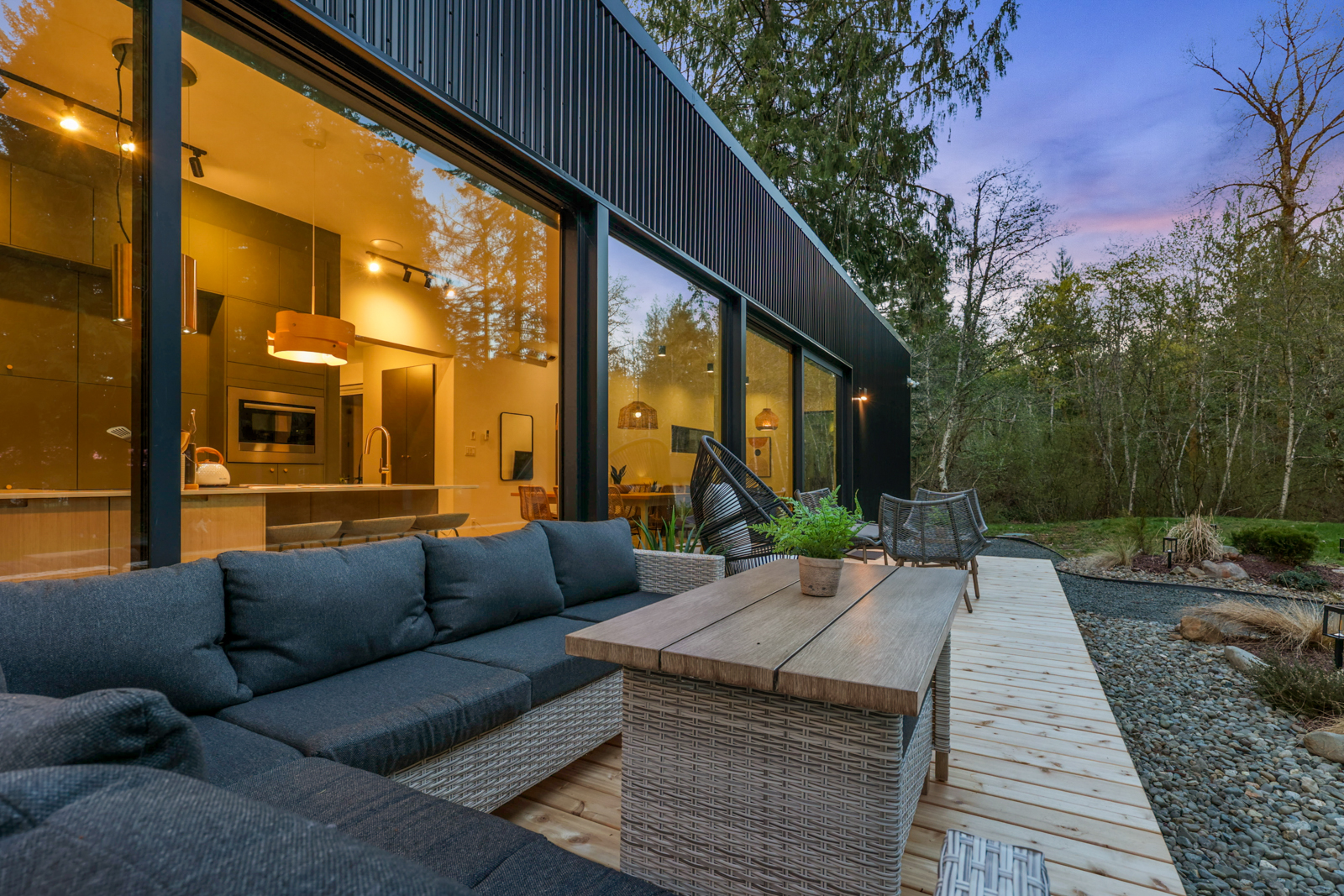 Main residence deck — outdoor sectional sofa, coffee table, and kitchen glowing through floor-to-ceiling glass at dusk
