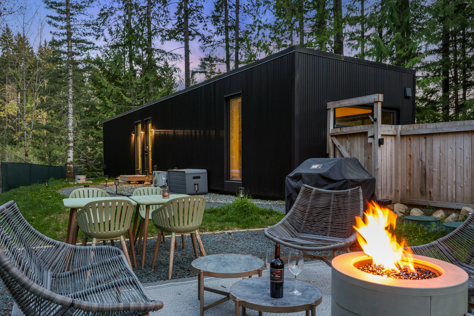 Guest house outdoor living — fire pit with flames, wine, dining table, and forest at dusk